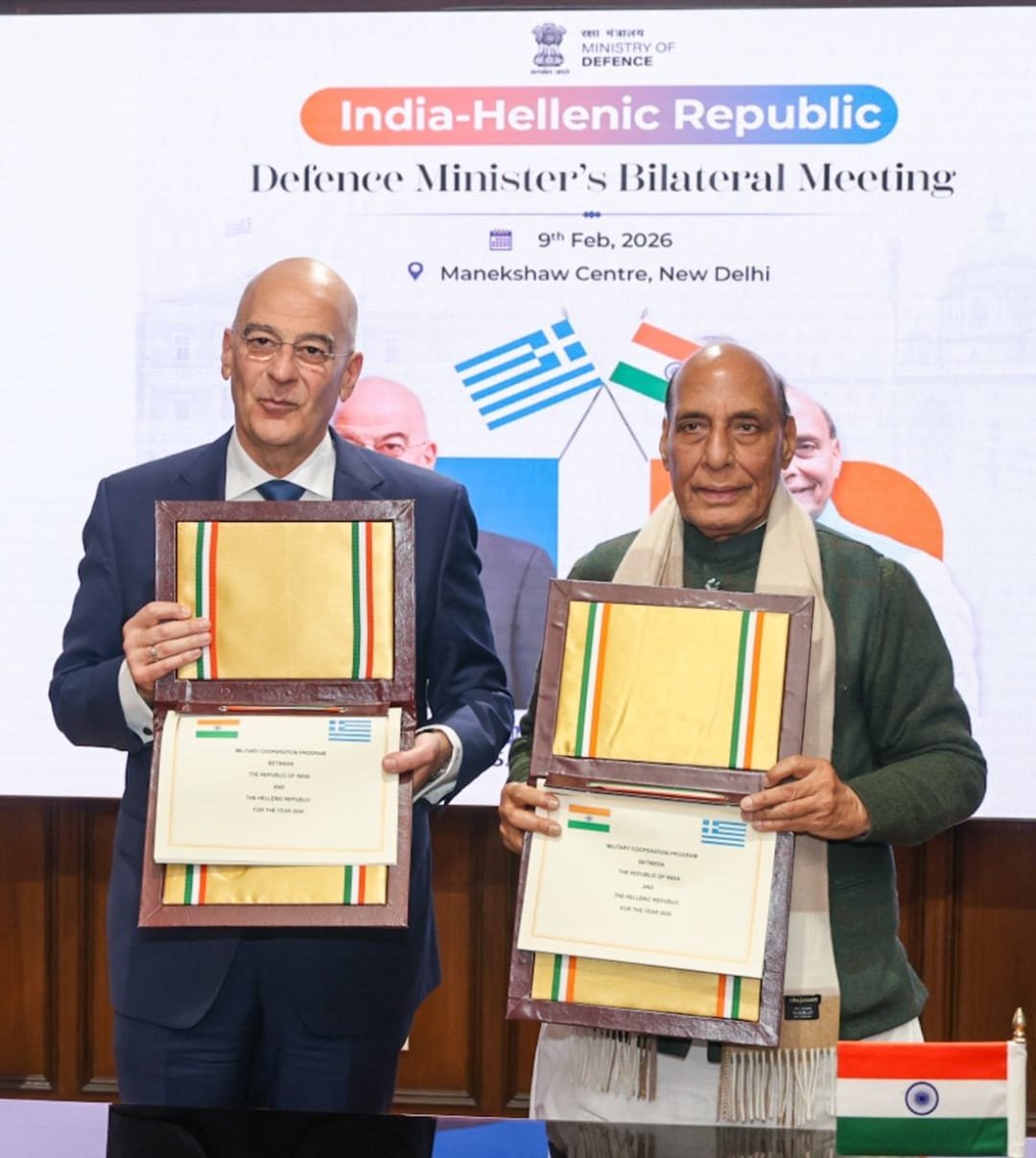 Two defense officials from india & greece hold signed documents during a bilateral defense meeting at the Manekshaw centre in New delhi , with the event backdrop and both national flags visible.